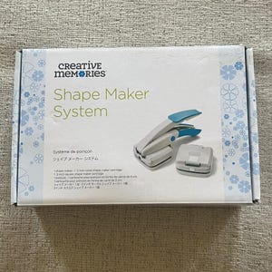 May include: A Creative Memories Shape Maker System box with a white and blue design. The box contains a white and blue shape maker tool and a square cartridge. The text on the box reads "Shape Maker System".