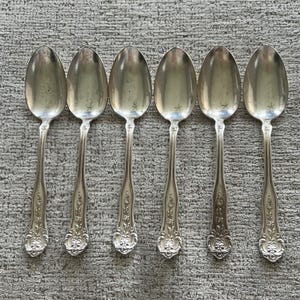May include: Six silver spoons with ornate floral detailing on the handles. The spoons have a classic, elegant design, with a polished finish. The spoons are arranged in a row, showcasing their symmetrical shape and reflective surfaces.