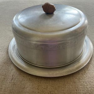 May include: A round, brushed aluminum cake dome with a matching circular tray base. The dome features a subtle embossed leaf pattern around its side and is topped with a brown, textured acorn-shaped handle made of a different material.