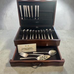 Könnte beinhalten: Eine dunkle Holzbesteckkiste, die geöffnet ist, um ein Set aus silbernen Utensilien zu zeigen. Das Set enthält Gabeln, Messer und Löffel, alle mit einem dekorativen Blumenmuster. Eine Schublade darunter enthält Servierteile und eine Garantiekarte.
