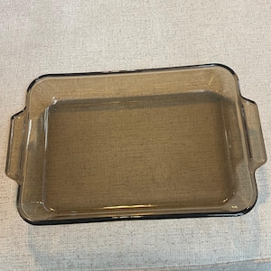 Puede incluir: Una fuente rectangular de vidrio transparente con esquinas redondeadas y dos asas. La fuente tiene un tinte ahumado marrón y está diseñada para uso en horno. La forma general es un diseño clásico para hornear y servir.
