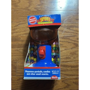 Op de afbeelding: Een blauw en bruin gumball-automaat met basketbalthema in de originele verpakking. De dispenser heeft een basketbalvormige bovenkant en een blauwe basis. De verpakking vermeldt "OFFICIAL SPORT Dispenser" en "INCLUDES GUMBALLS". Het geeft gumballs, snoep en andere kleine snacks af.