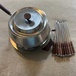 May include: A stainless steel fondue pot with a dark wood handle and lid knob. A set of fondue forks with wooden handles are arranged next to the pot. The pot sits on a round base.