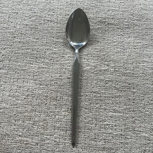 Könnte beinhalten: Ein Edelstahl-Löffel mit einem langen, schlanken Stiel und einer ovalen Schüssel. Der Griff hat ein facettiertes Design in der Nähe der Schüssel. Der Löffel ist vor einem strukturierten, grauen Hintergrund platziert.