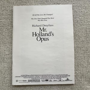 Op de afbeelding: Een wit papier met de titel "Mr. Holland's Opus" en de naam Richard Dreyfuss. De tekst "Of All The Lives He Changed, The One That Changed The Most Was His Own" is ook zichtbaar.