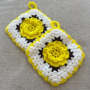 Peut inclure: Deux dessous de plat faits à la main au crochet. Chaque dessous de plat présente un motif de fleur jaune au centre, entouré de carrés blancs et marron, avec une bordure jaune. Une petite boucle jaune est attachée pour suspendre.