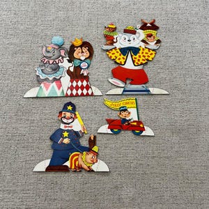Puede incluir: Una colección de recortes de papel vintage con temática de circo. El conjunto incluye un elefante, un león, un oso, un payaso y un coche con una pancarta que dice "JOLLY CIRCUS". Cada recorte es de colores brillantes y presenta detalles intrincados.