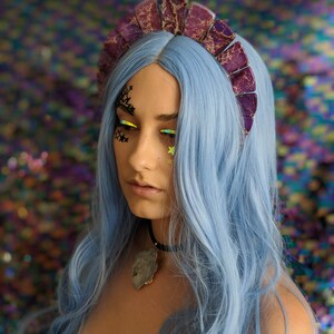 May include: A woman with blue hair is wearing a purple and silver crown with a jagged edge. She is wearing a black choker with a large, gray, crystal pendant.