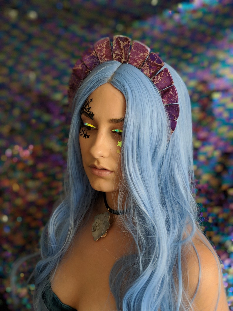 May include: A woman with blue hair is wearing a purple and silver crown with a jagged edge. She is wearing a black choker with a large, gray, crystal pendant.