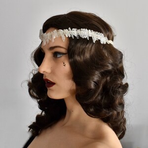 May include: A woman with dark brown hair styled in loose curls wears a white crystal hair crown. The crown is made of many small, pointed crystals.