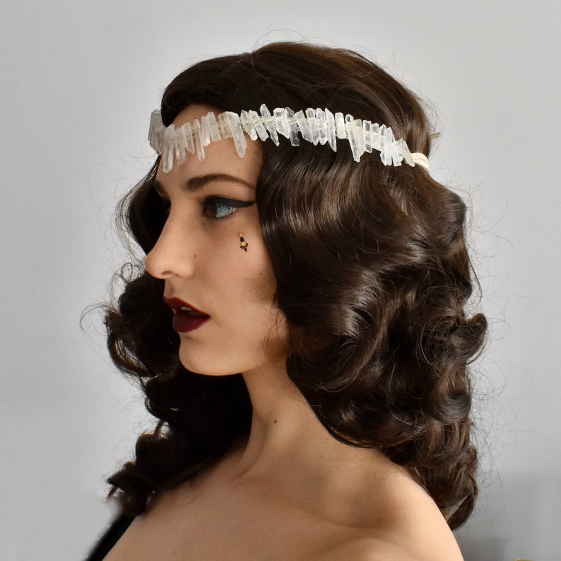 May include: A woman with dark brown hair styled in loose curls wears a white crystal hair crown. The crown is made of many small, pointed crystals.