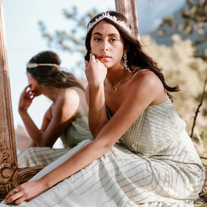 May include: A woman wearing a white crystal crown and a light green sequined dress sits in front of a mirror. She is looking at the camera with a serious expression.