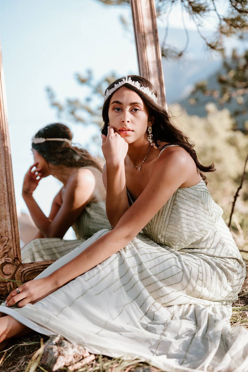 May include: A woman wearing a white crystal crown and a light green sequined dress sits in front of a mirror. She is looking at the camera with a serious expression.