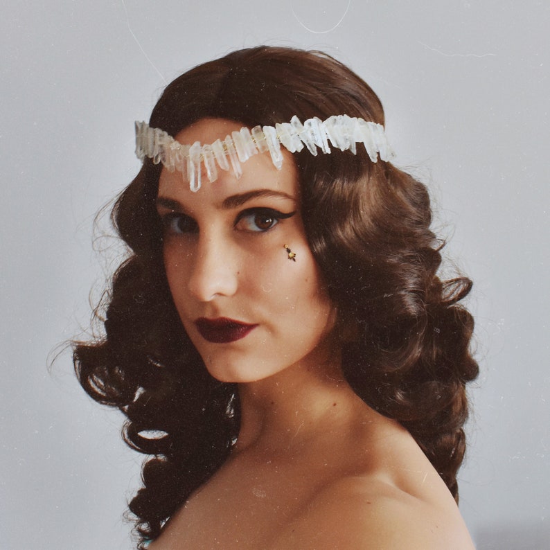 May include: A woman with dark brown curly hair wears a white crystal crown. She has dark eye makeup and dark red lipstick.