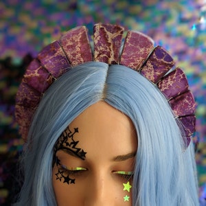 May include: A purple and gold marbled headband with a scalloped edge. The headband is worn by a person with blue hair and black star-shaped makeup around their eyes.