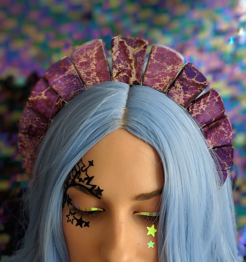 May include: A purple and gold marbled headband with a scalloped edge. The headband is worn by a person with blue hair and black star-shaped makeup around their eyes.
