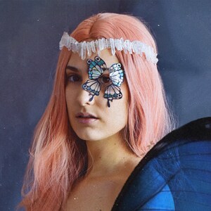 May include: A woman with pink hair wears a crystal headband and a butterfly costume. The butterfly wings are blue and black. The butterfly costume has a butterfly design on the woman's face.
