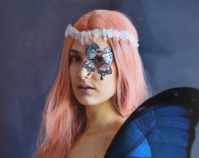 May include: A woman with pink hair wears a crystal headband and a butterfly costume. The butterfly wings are blue and black. The butterfly costume has a butterfly design on the woman's face.