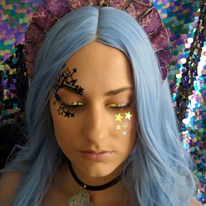 May include: A woman with blue hair is wearing a purple and white crown with a black choker necklace with a large grey and white stone pendant. She has black star-shaped stickers on her face and gold star-shaped stickers on her cheek.