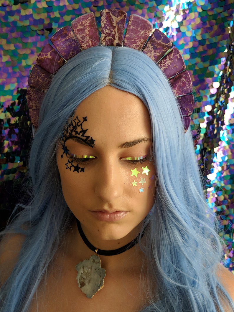 May include: A woman with blue hair is wearing a purple and white crown with a black choker necklace with a large grey and white stone pendant. She has black star-shaped stickers on her face and gold star-shaped stickers on her cheek.