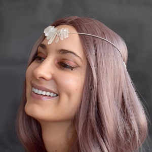 May include: A woman with long, pink hair is wearing a crystal headband. The headband is made of a thin silver wire with clear quartz crystals attached to it. The crystals are arranged in a fan shape and are pointing upwards. The woman is smiling and looking to the side.