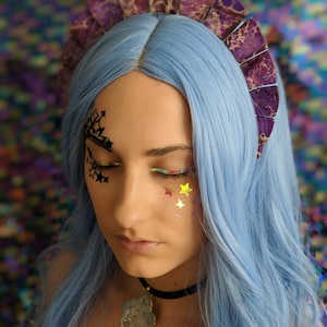 May include: A woman with blue hair is wearing a purple and white crown with a large, multi-colored stone pendant necklace. She has glitter stars on her face.