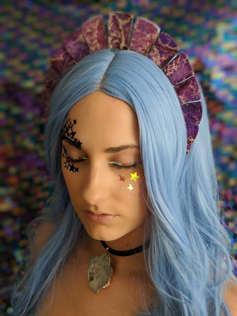 May include: A woman with blue hair is wearing a purple and white crown with a large, multi-colored stone pendant necklace. She has glitter stars on her face.