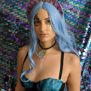 May include: A woman with blue hair and a purple crown is wearing a blue and black swimsuit with a seashell design. She is wearing a necklace with a large, clear crystal pendant.