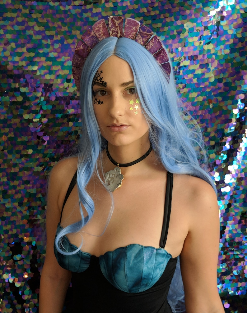 May include: A woman with blue hair and a purple crown is wearing a blue and black swimsuit with a seashell design. She is wearing a necklace with a large, clear crystal pendant.
