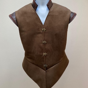 以下が含まれることがあります： 茶色のスエードベストで、先のとがった裾と茶色の革のトリムが付いています。ベストはハイカラーで、前面に真鍮製のボタンが5つ付いています。