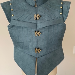 May include: A blue-grey sleeveless vest featuring a high collar, vertical and diagonal quilted stitching, and three antique brass-toned metal toggle clasps down the center front. The shoulders are accented with small, round brass-toned metal studs.
