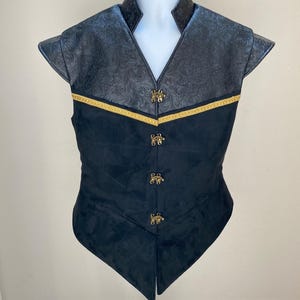 May include: A black vest featuring a textured floral pattern on the upper chest and shoulders, separated from the solid black lower section by a gold braided trim. The garment includes four ornate gold-toned metal clasps down the front center.
