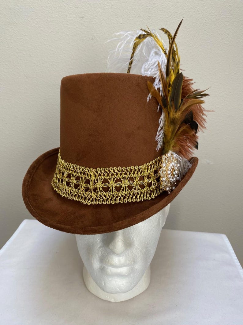 May include: A brown top hat with a gold filigree band and decorative feathers. The hat features a cluster of brown, white, and yellow feathers and a jeweled accent. This formal hat is suitable for costume or special events.