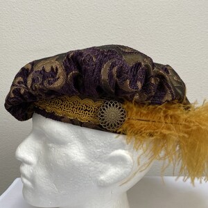 May include: A purple and gold patterned beret with a gold trim and a gold feather accent. The beret is worn on a white mannequin head.