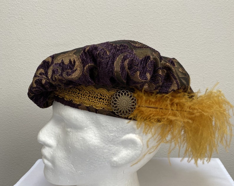 May include: A purple and gold patterned beret with a gold trim and a gold feather accent. The beret is worn on a white mannequin head.