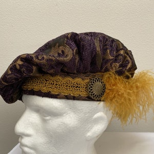 May include: A purple velvet beret with a gold trim and a gold feather accent. The beret has a patterned design.