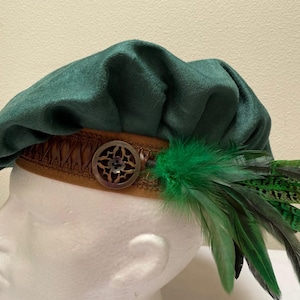 May include: A dark green velvet beret featuring a tan band with a decorative brown circular metal button and a cluster of green and brown patterned feathers attached to the side. The hat is displayed on a white mannequin head form.