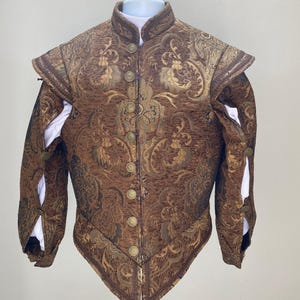 May include: A brown and gold brocade jacket with a high collar and decorative buttons. The jacket features a detailed floral pattern and white fabric peeking from the sleeves. It is likely a historical costume or theatrical garment.