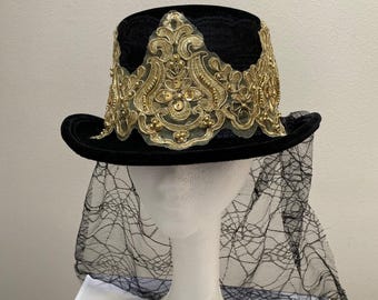 May include: Black top hat with a gold embroidered design and a black lace veil. The hat features a velvet crown and brim, with the gold embroidery adding a decorative element. The veil is attached to the hat and drapes down.