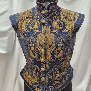 Puede incluir: Un chaleco azul y dorado de brocado con cuello alto y mangas cortas. El chaleco tiene un patrón decorativo y se cierra con botones dorados.