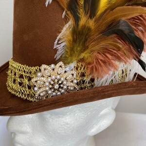May include: A brown top hat with a gold filigree band and a jeweled floral embellishment. The hat is adorned with colorful feathers in shades of yellow, brown, and white, creating a striking visual contrast.