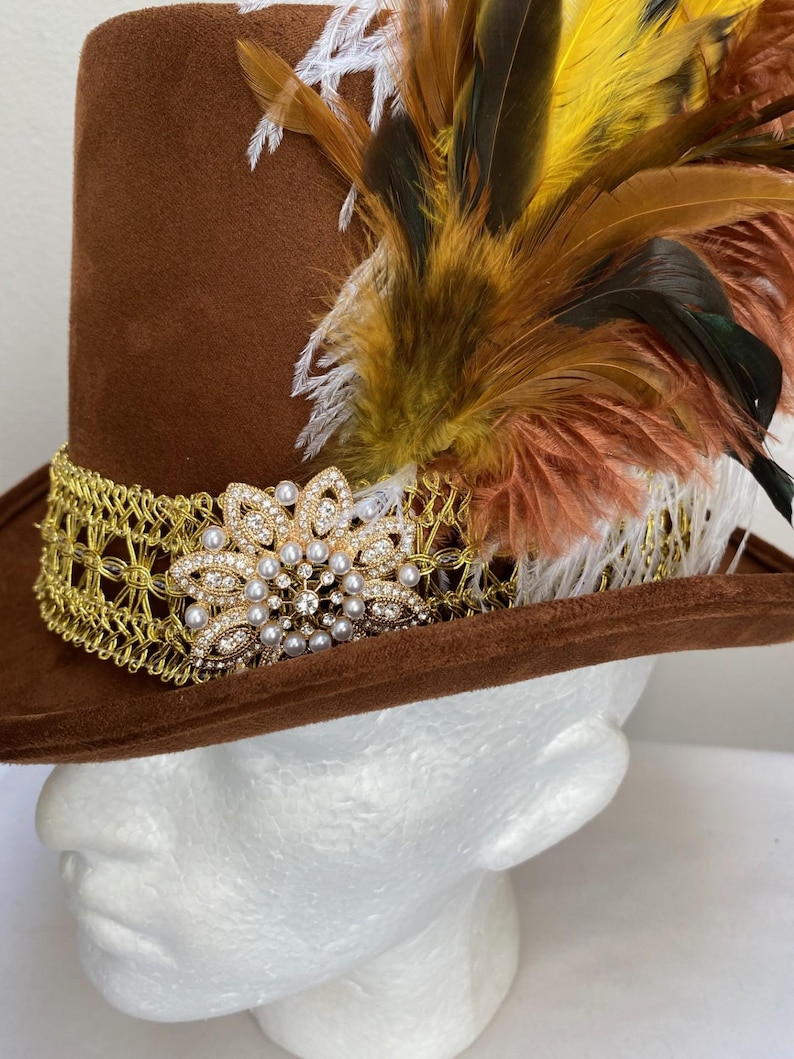 May include: A brown top hat with a gold filigree band and a jeweled floral embellishment. The hat is adorned with colorful feathers in shades of yellow, brown, and white, creating a striking visual contrast.