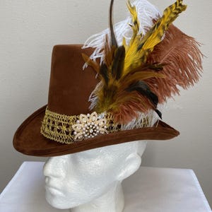 May include: A brown suede hat with a wide brim, adorned with a gold lace band and a sparkling rhinestone and pearl embellishment. The hat is decorated with a cluster of feathers in shades of brown, yellow, and white.