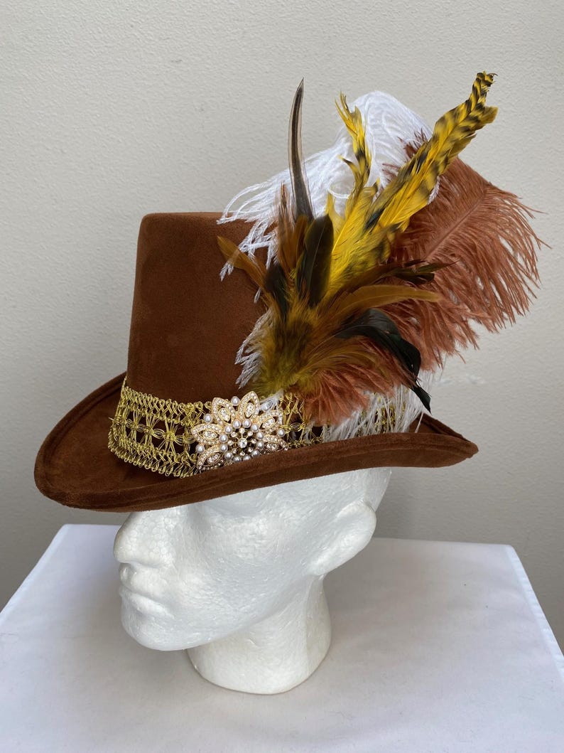 May include: A brown suede hat with a wide brim, adorned with a gold lace band and a sparkling rhinestone and pearl embellishment. The hat is decorated with a cluster of feathers in shades of brown, yellow, and white.