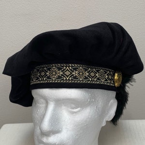 Op de afbeelding: Een zwarte fluwelen baret met een gouden en zwarte band met patroon en een zwarte verenversiering.