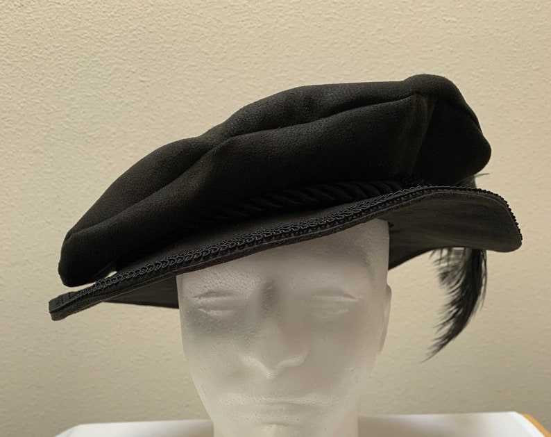 May include: A black felt hat with a black cord trim and a black feather. The hat has a slightly rounded crown and a wide brim.
