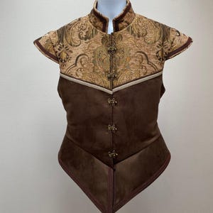Puede incluir: Un chaleco marrón con cuello alto y mangas cortas. La parte superior presenta un estampado de brocado floral dorado y marrón, mientras que la sección inferior es de material liso marrón similar al ante. El chaleco tiene cierres y adornos decorativos.