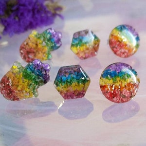 May include: Six rainbow-coloured resin cabochons with embedded dried flowers. The cabochons are in various shapes, including a circle, a hexagon, and a hand-shaped design.