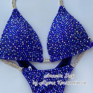 Puede incluir: Un bikini azul real con adornos de cristal. La parte superior del bikini tiene un estilo halter y la parte inferior es de corte alto. El bikini está adornado con diamantes de imitación transparentes y cristales iridiscentes. El texto "Amnesia Shop by Alyona Konstantinova" está impreso en la parte inferior del bikini.