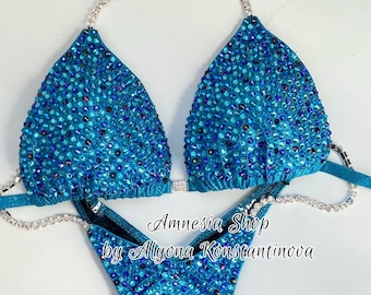 May include: Teal blue bikini with crystal embellishments. The bikini top has a halter style with a silver chain strap. The bikini bottom has a high-cut style. The bikini is labeled "Amnesia Shop by Allyona Konstantinova".
